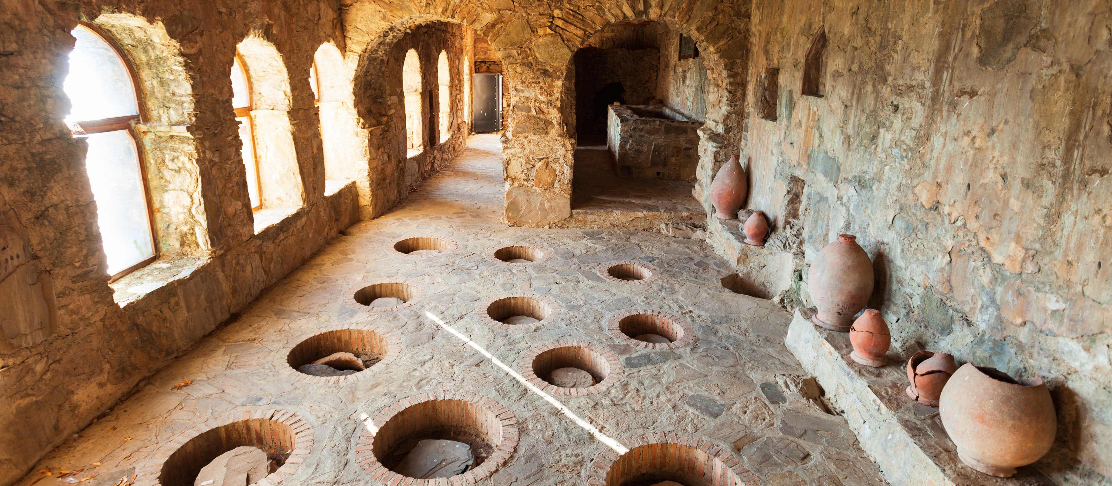 Traditional Georgian winemaker working with qvevri clay vessels in ancient wine cellar with stone walls and wooden tools