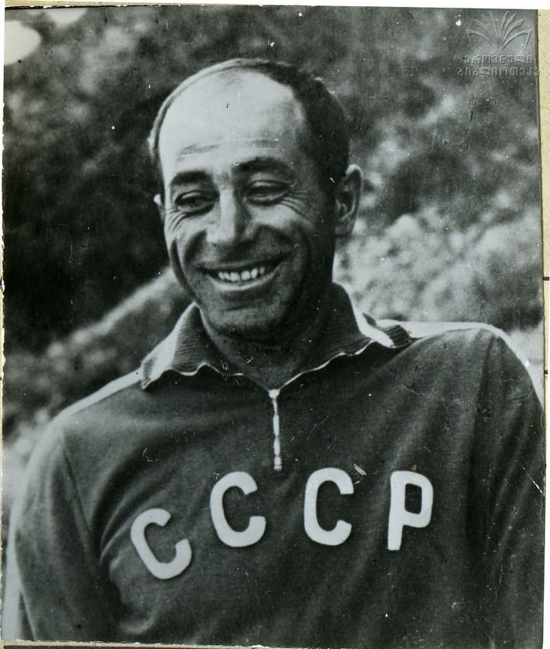 Black-and-white archive portrait of a smiling mountaineer wearing a Soviet team jersey with the letters CCCP across the chest, trees behind him