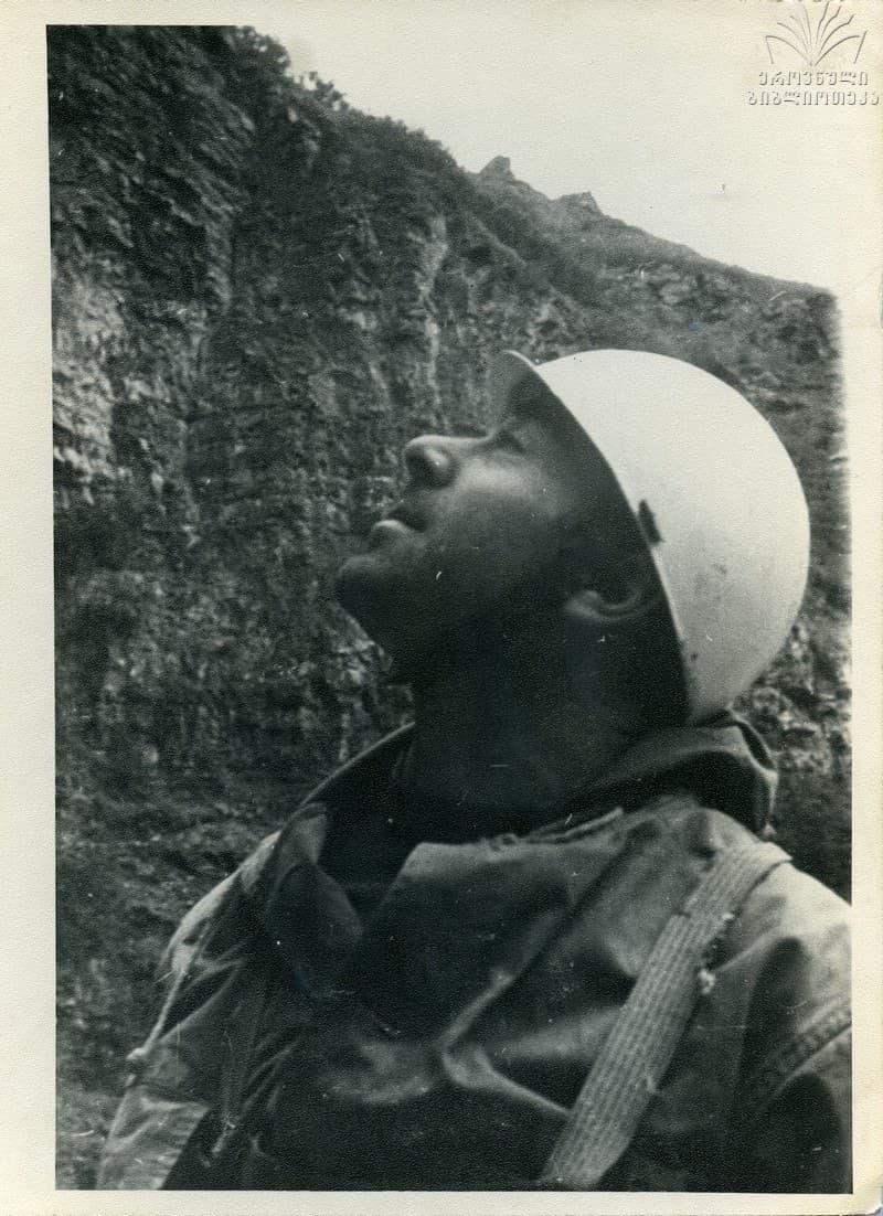Black-and-white archive photograph of a climber in a helmet and heavy jacket looking up at a steep, layered rock face above him