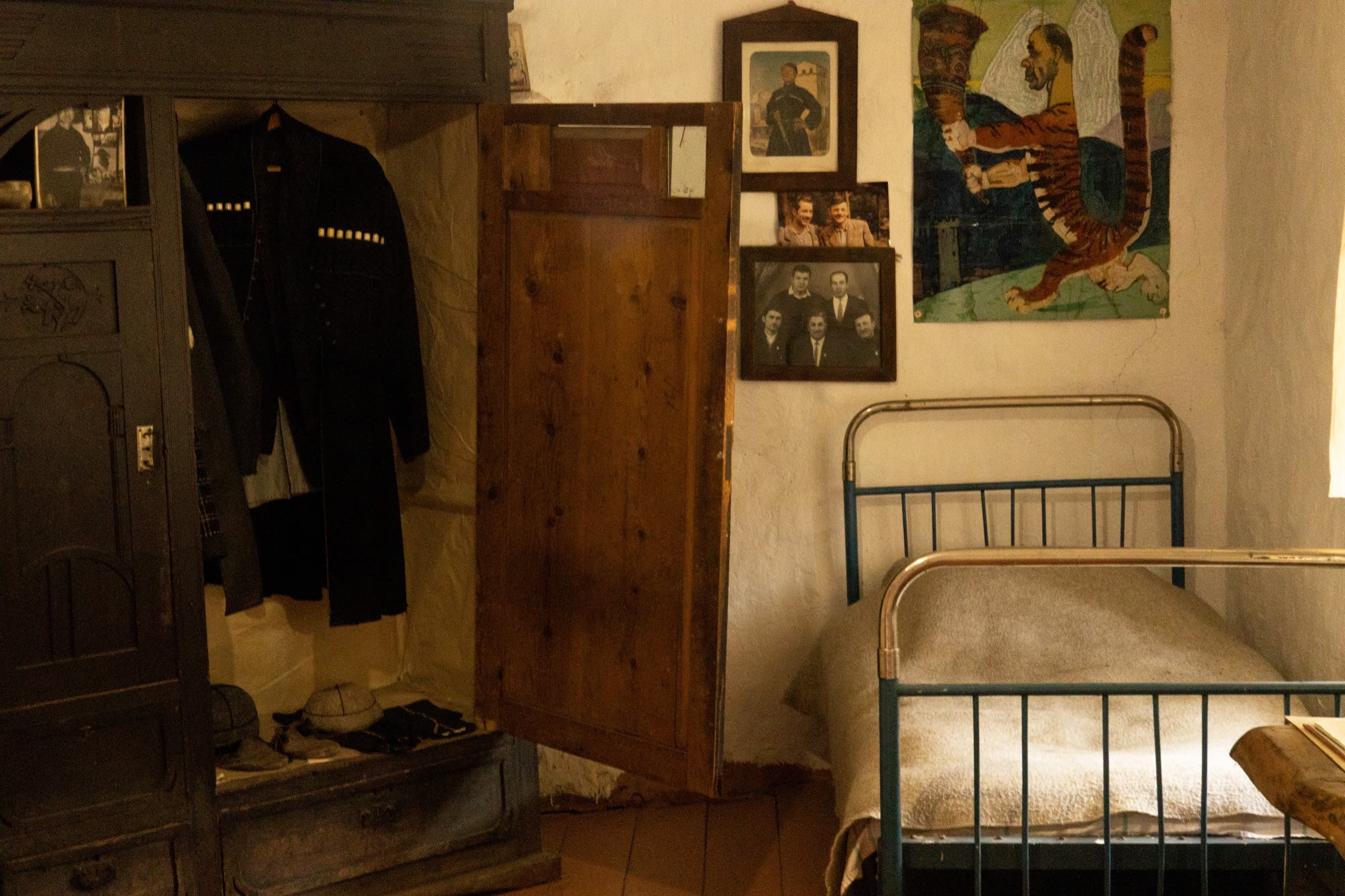 Interior of the Khergiani house-museum in Mestia: a dark wooden wardrobe holding a jacket, an iron bedstead, framed family photographs on the whitewashed wall and a painted panel of a tiger
