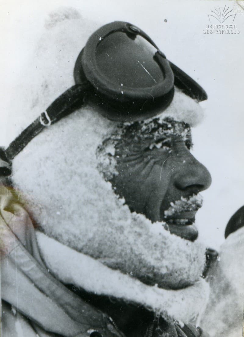 Black-and-white archive photograph of a mountaineer's face crusted with snow, wearing a hooded jacket with glacier goggles pushed up onto his forehead