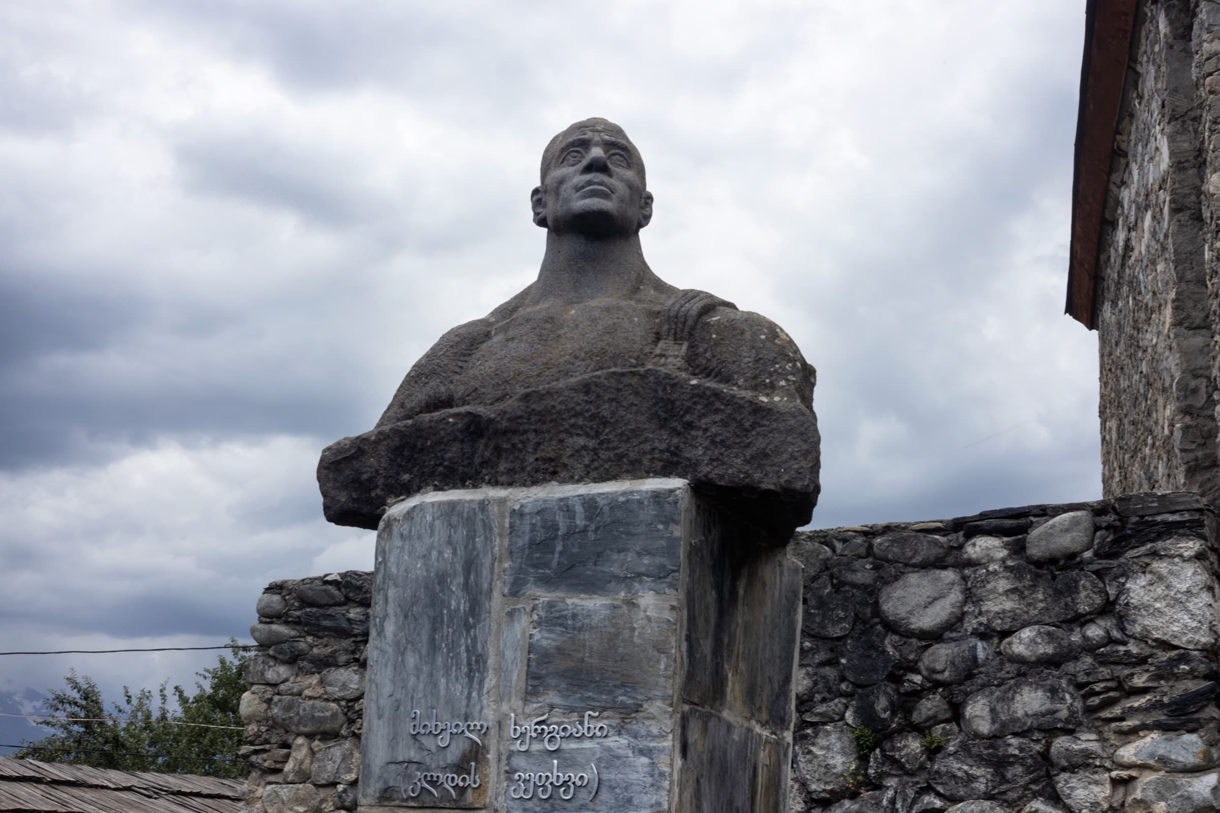 Mikheil Khergiani - the Tiger of the Rocks from Svaneti Stone bust of the Georgian mountaineer Mikheil Khergiani on a plinth in Mestia, backed by a dry-stone wall under heavy grey cloud