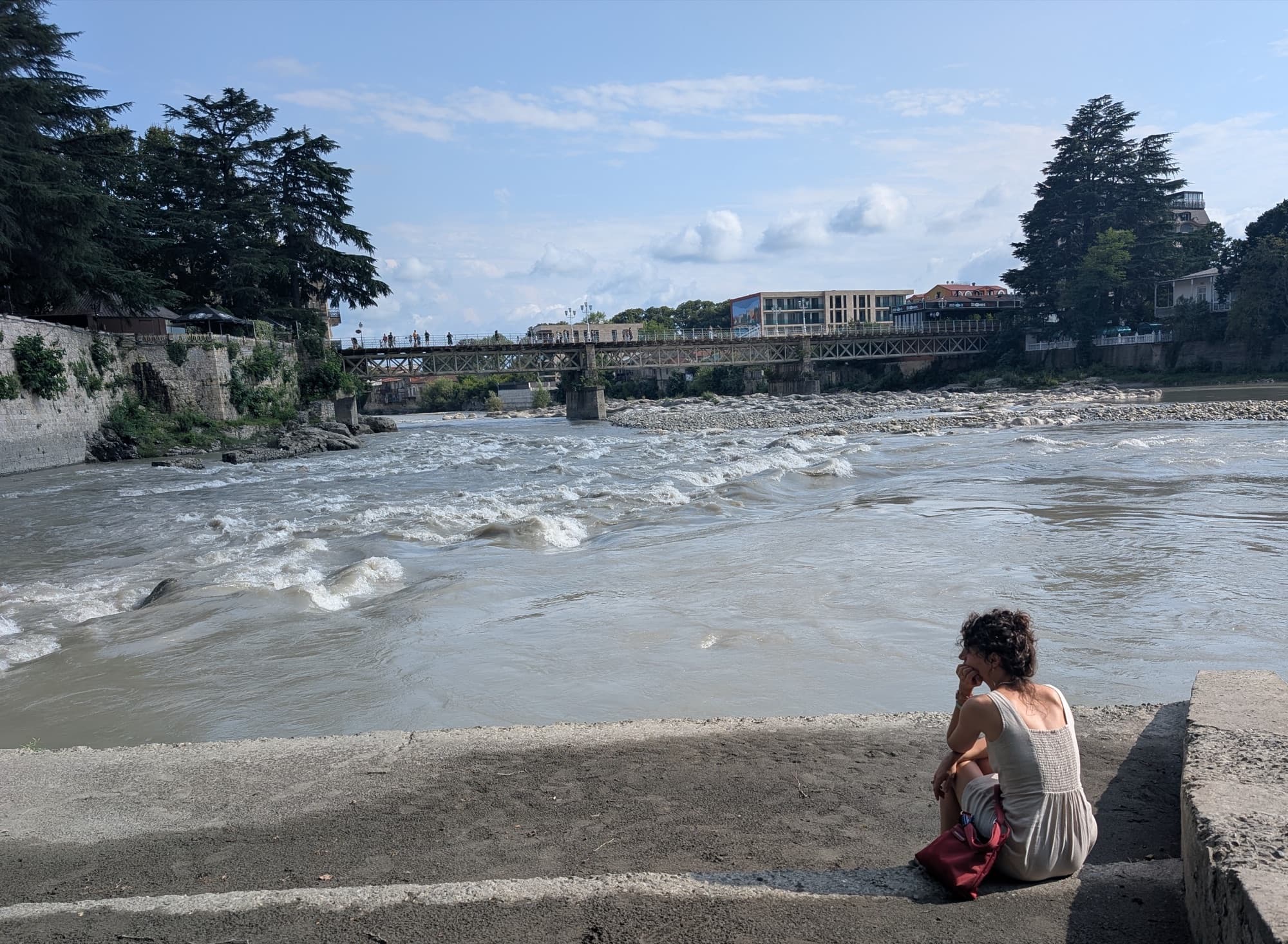 A woman sitting on stone steps above the Rioni river in Kutaisi, looking toward a bridge