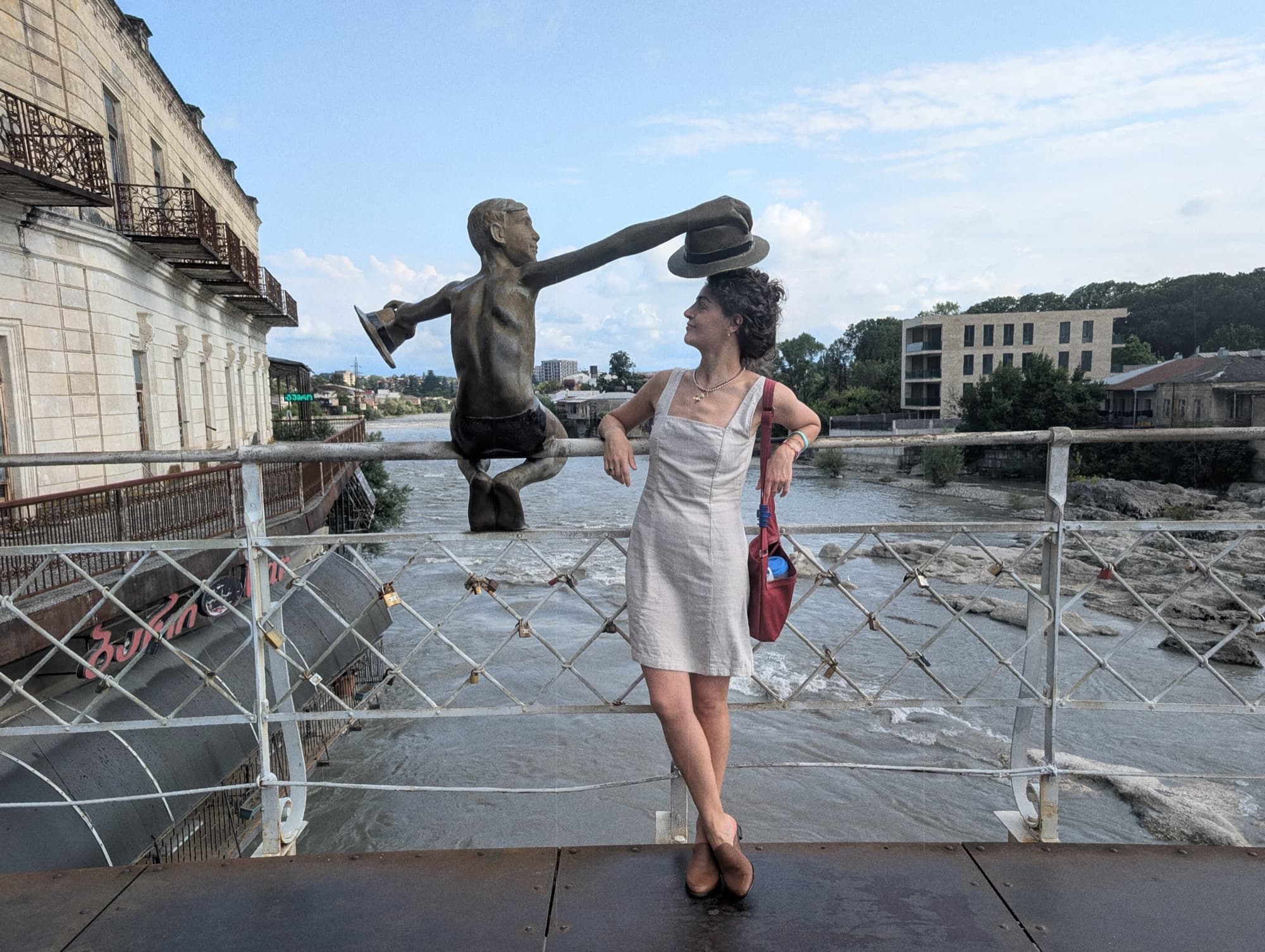 A woman standing on Kutaisi's White Bridge beside the bronze statue of a boy about to jump into the Rioni river