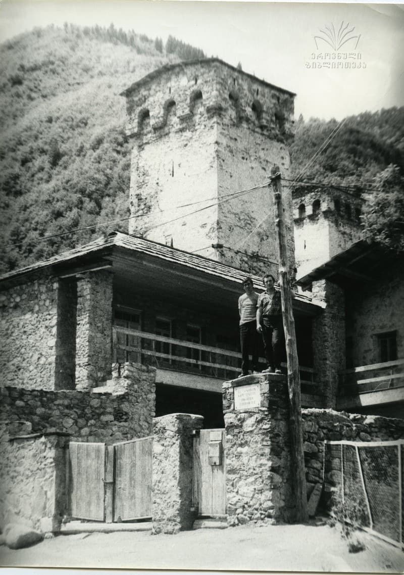 Black-and-white archive photograph of a Svan defensive tower rising behind a stone house with a wooden balcony in Mestia, two young men standing on the gatepost below