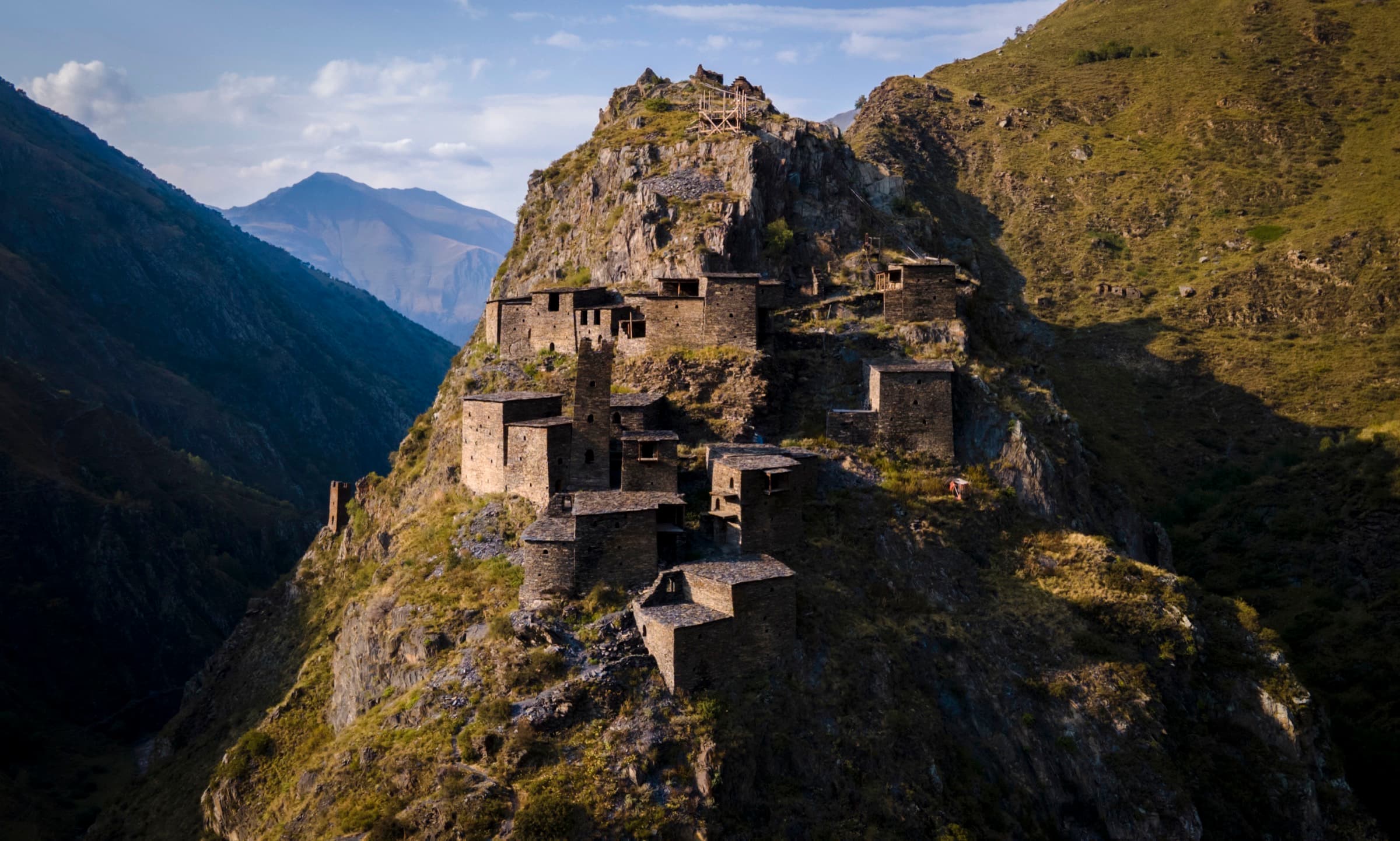 Aerial view of Mutso fortress-village's stone towers perched on a rocky cliff outcrop in Khevsureti, Georgia, lit by golden afternoon light