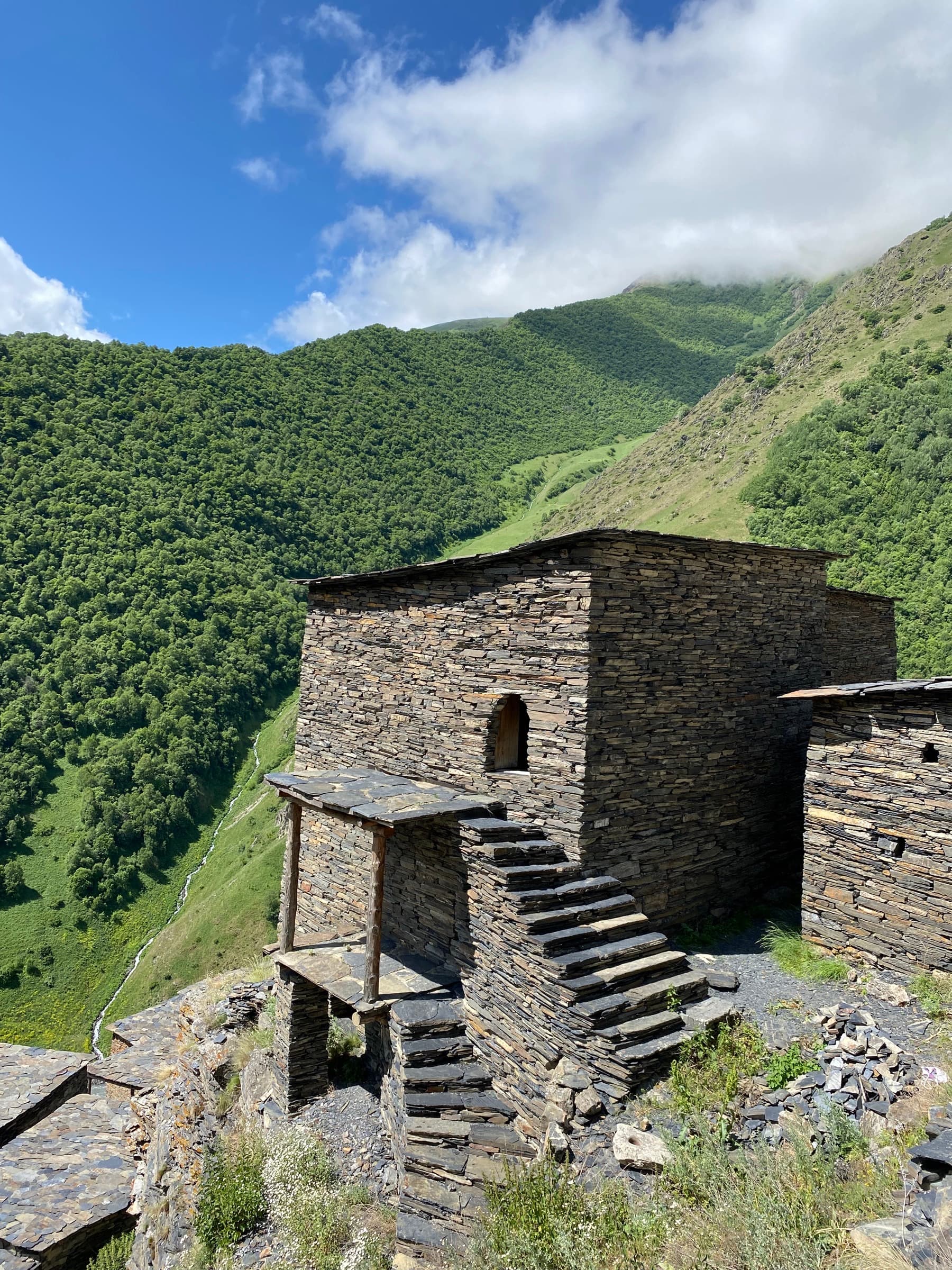A multi-story fortified residence in Mutso built from stacked slate stone, with a slate staircase leading up to a small arched doorway, perched on a cliff edge above a green mountain valley