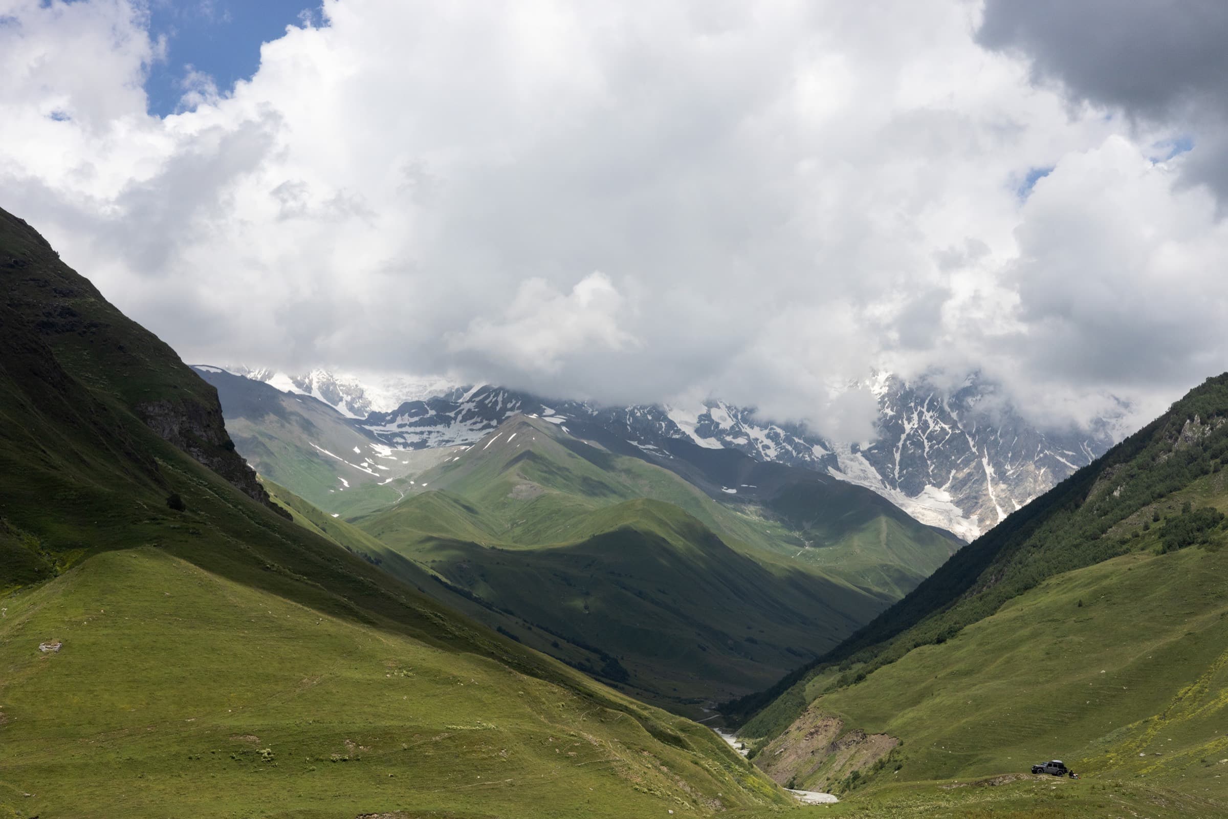 A green Svaneti valley in summer with a river running through it and the glaciated peaks of the Caucasus range half-hidden in cloud at its head